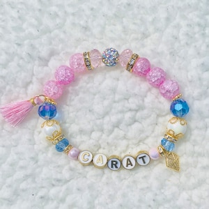 May include: A pink and blue beaded bracelet with a gold charm that says "CARAT". The bracelet has a pink tassel and a clear crystal bead.