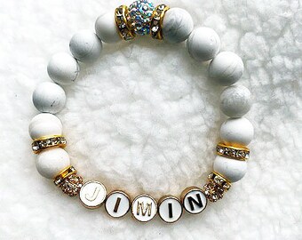 May include: White beaded bracelet with gold accents and a crystal charm. The bracelet has the name "Jimin" spelled out in gold letters.
