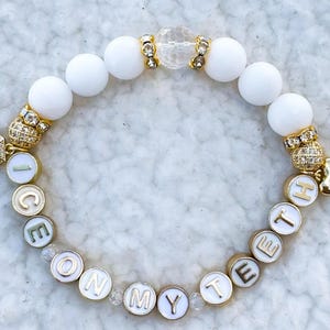 May include: White beaded bracelet with gold charms spelling out "ICE ON MY TEETH" and a gold tooth charm. The bracelet also features a clear crystal charm.
