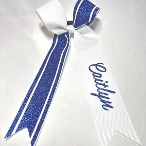 May include: A cheer bow featuring white and blue glitter ribbon with a central knot. One long tail displays the name Caitlyn in blue glitter script, while the other tail features blue glitter stripes bordered by thin white lines on a white base.