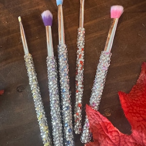 May include: Five makeup brushes with silver handles encrusted with rhinestones. The brush heads are various colors, including yellow, purple, blue, orange, and pink. The brushes are arranged on a dark wooden surface.