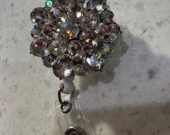 May include: A sparkly badge reel with a cluster of clear, faceted rhinestones. The rhinestones are arranged in a floral pattern and have iridescent highlights. The reel has a clear plastic strap and a metal clip.