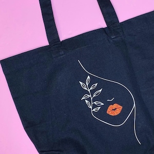 May include: Black tote bag with a white line drawing of a face with a single leaf branch and red glitter lips.