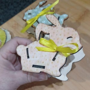 May include: Three wooden bunny shaped ornaments with yellow ribbon bows. The bunnies are painted with a light brown base and white polka dots.