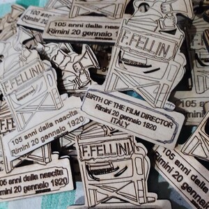 May include: A collection of wooden plaques with a black and white illustration of a man holding a megaphone. The plaques commemorate the 105th anniversary of the birth of Federico Fellini, an Italian film director, in Rimini, Italy. The text on the plaques reads "105 anni dalla nascita Rimini 20 gennaio 1920" and "Birth of the Film Director Rimini 20 gennaio 1920 Italy".