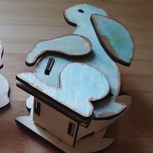 Può includere: Una statuetta di coniglio di legno blu chiaro con una finitura di vernice invecchiata. Il coniglio è seduto su una base di legno con un piccolo design di casa.