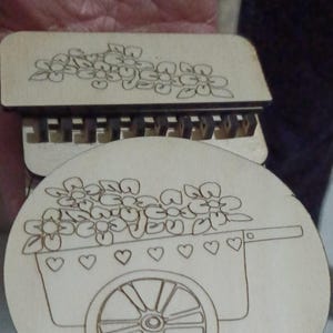 Op de afbeelding: Een houten uitsnijding van een bloemenkar met een wiel en bloemen in de kar. De kar heeft hartjes aan de zijkant en de tekst "by Vivedo3D" staat onderaan.