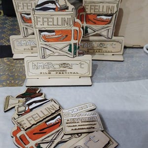 May include: Wooden award for the Amarcort Film Festival. The award features a cartoon director holding a megaphone and sitting in a director's chair. The award is inscribed with the text "F. Fellini" and "Amarcort Concorso Film Festival".