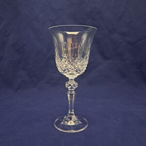 May include: A clear crystal wine glass with a diamond-cut pattern on the bowl. The stem features a decorative, faceted design. The glass sits on a round base, and the background is a solid dark blue.
