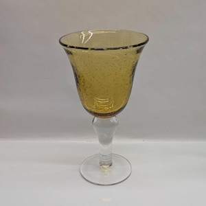 May include: A single amber-colored wine glass with a clear stem and base. The glass has a flared bowl with small bubbles throughout the glass. The rim of the glass is dark amber.