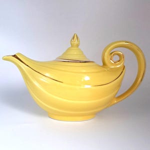 May include: A bright yellow ceramic teapot featuring a curved handle, a pointed spout, and a matching lid with a finial. The body of the teapot is decorated with horizontal ridges and thin gold trim accents along the spout, lid, and handle edges.