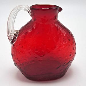 Könnte beinhalten: Ein runder, rubinroter Glaskrug mit einer texturierten, genoppten Oberfläche und einem klaren, gerippten Glasgriff. Das Gefäß hat einen breiten, bauchigen Körper, der sich zu einer kleinen, offenen Ausgussöffnung an der Oberseite hin verjüngt.