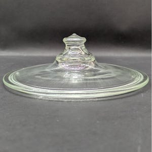 May include: A clear, circular glass lid featuring a smooth, flat rim and a central, tiered knob handle. The lid is transparent, allowing light to pass through its surface, and it has a slightly raised, rounded top section for easy gripping.