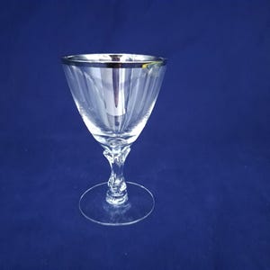 Peut inclure: Un verre transparent avec un bord argenté, présentant une coupe conique avec des rainures verticales et une tige décorative. Le verre repose sur une base plate et circulaire. Le fond est bleu foncé. Idéal pour servir des boissons.