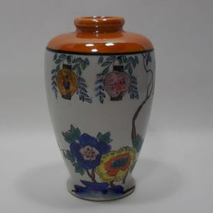 Puede incluir: Un jarrón de cerámica con cuerpo blanco y borde naranja. El jarrón está decorado con coloridos diseños florales y de linternas. Las flores son azules, amarillas y naranjas. Las linternas son amarillas y rosas.