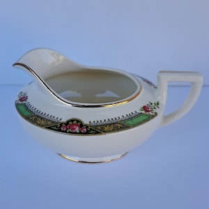 May include: A cream-colored ceramic creamer featuring a gold-trimmed rim and handle. The body is decorated with a band of black, green, and gold geometric patterns interspersed with small pink rose floral motifs and delicate white dot accents.