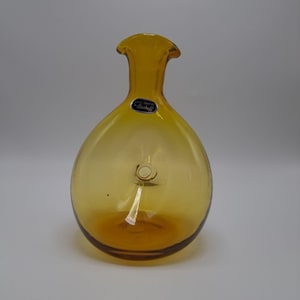May include: A vintage amber-colored glass vase with a rounded body and a flared neck. The vase has a label that reads "Hand Crafted Bischoff". The glass has a glossy finish and a slight gradient in color.