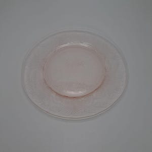 Puede incluir: Un plato de cristal transparente y rosa con un diseño floral. El plato tiene un centro circular elevado y un borde exterior más ancho, ambos decorados con flores y hojas en relieve. El plato es probablemente para servir o exhibir.