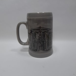 May include: Gray ceramic beer stein with a handle, featuring a detailed black and white illustration of a building facade. The stein has a cylindrical shape and a slightly flared base. The handle is curved and attached to the side of the stein.