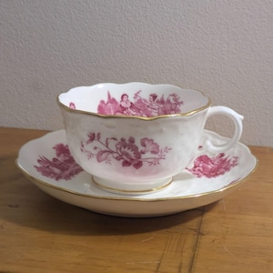 May include: A white teacup and saucer set with a pink floral and scenic design. The cup and saucer have a gold trim. The cup has a handle and a scalloped edge. The saucer is round with a matching design.