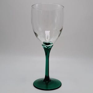 May include: A clear glass wine goblet featuring a smooth, transparent bowl that transitions into a deep emerald green stem and circular base. The stem has a distinct, faceted, leaf-like design where it meets the bottom of the glass bowl.