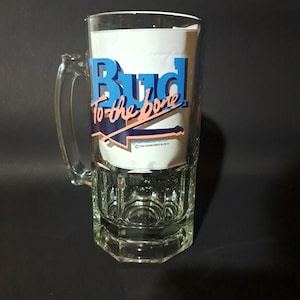 May include: Clear glass beer mug with a handle. The mug features the Budweiser logo in blue and pink, with the phrase "Bud to the bone." The mug has a rectangular base and a clear handle.