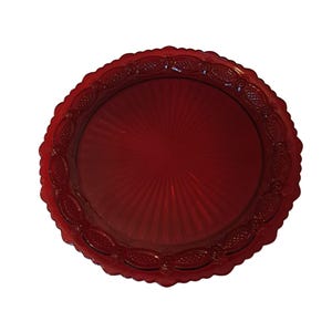 May include: A circular ruby red glass plate featuring a scalloped edge and a decorative border with embossed oval and diamond patterns. The center of the plate displays a radiating sunburst design that extends outward from the middle point.