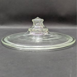 May include: A clear glass lid with a knob handle. The lid is circular with a slightly raised edge. The knob is a small, detailed design. The glass is transparent, allowing a view of the background.