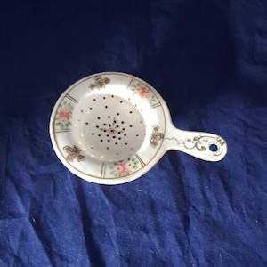 Puede incluir: Colador de té de porcelana blanca con asa, con rosas rosas y detalles florales dorados. El colador tiene un cuenco circular perforado y un asa decorativa con un agujero. Diseño vintage y elegante.