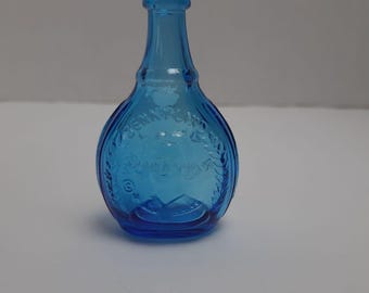 Peut inclure: Petite bouteille en verre bleu avec un corps arrondi et un long col. La bouteille présente du texte en relief et des motifs floraux. La bouteille est vide et repose sur une surface blanche. La bouteille mesure environ 13 cm de haut.