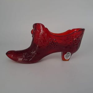 Puede incluir: Un zapato de cristal rojo rubí con detalles florales. El zapato decorativo tiene una punta puntiaguda y un tacón pequeño. El cristal es translúcido y el zapato es probablemente un artículo de colección o decorativo.