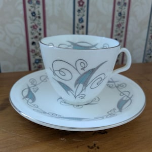 May include: A white teacup and saucer set with a silver trim, featuring a design of blue and grey leaf-like shapes and swirls. The teacup has a handle and sits on a matching saucer. The set is on a wooden surface.