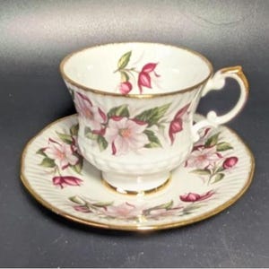 Op de afbeelding: Een witte theekop en schotelset met een bloemenontwerp. De kop en schotel zijn versierd met roze en rode bloemen en groene bladeren. De randen en het handvat zijn goudkleurig afgewerkt. De schotel heeft een geribbeld patroon.