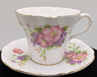 May include: White teacup and saucer set with a floral design featuring pink and purple flowers and green leaves. The cup and saucer have gold trim. The teacup has a curved handle and a scalloped rim. The saucer is also scalloped.