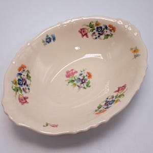 Puede incluir: Un cuenco ovalado de cerámica de color crema con un borde festoneado. El interior presenta un ramo floral central y cuatro grupos florales más pequeños alrededor del borde, con flores rosas, azules, amarillas y naranjas con hojas verdes.