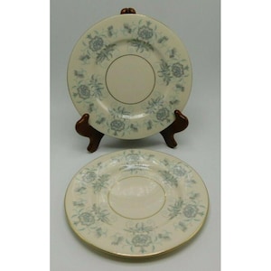 May include: Two cream-colored plates with a floral pattern in shades of blue and a gold rim. The plates are displayed on a wooden stand. The plates have a central cream-colored circle.