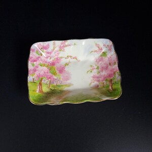 May include: A rectangular, ceramic dish with a hand-painted scene of pink cherry blossom trees lining a path. The dish has a gold trim and a slightly scalloped edge. The background is a soft white.