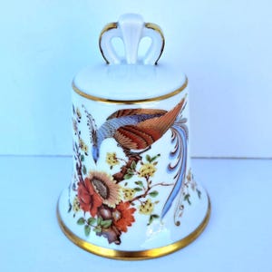 May include: A white porcelain bell featuring a hand-painted design of a long-tailed bird perched on a branch with orange and yellow flowers. The bell has a gold-trimmed rim and a decorative arched handle at the top, finished with gold accents.