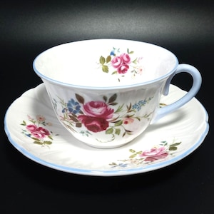 以下が含まれることがあります： ピンク、青、緑の色合いのバラの花柄が施された、白のティーカップとソーサーのセット。カップは水色の持ち手と縁、ソーサーはそれに合わせた青い縁取り。お茶用です。