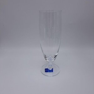 May include: A clear, stemmed champagne flute with a round base. The glass has a tall, slender bowl and a small, circular foot. The glass is made of clear glass and is empty. A blue sticker is on the base.