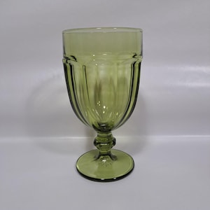 May include: A green-tinted glass goblet with a ribbed design on the bowl. The goblet has a round base and a decorative stem. The glass is transparent, allowing light to pass through, and is suitable for serving beverages.