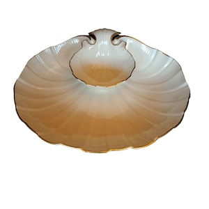 May include: A white and gold seashell-shaped serving dish. The dish has a large, scalloped outer section and a smaller, circular inner section. The edges are trimmed with gold. The dish is designed to hold food items.