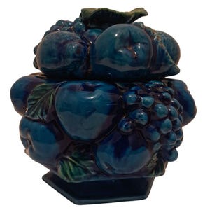 May include: A dark blue glazed ceramic jar with a lid, featuring a three-dimensional relief design of various fruits including apples and grapes. The object has a glossy finish and sits on a hexagonal base, showcasing deep blue and green tones.
