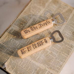 May include: Two wooden bottle openers with stainless steel tops. One opener has "We The People" engraved on the handle. The other has coordinates 27° 20' 39.404" N 80° 21' 19.393" W. The openers rest on a vintage newspaper.