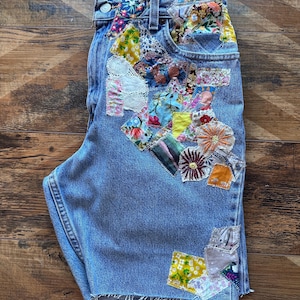 May include: Light blue denim shorts with a patchwork design featuring various floral and geometric patterns. The patchwork is sewn onto the front of the shorts, covering the front pocket and extending down the side seam.