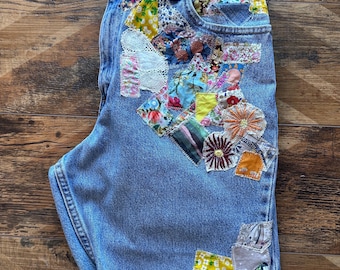 May include: Light blue denim shorts with a patchwork design featuring various floral and geometric patterns. The patchwork is sewn onto the front of the shorts, covering the front pocket and extending down the side seam.