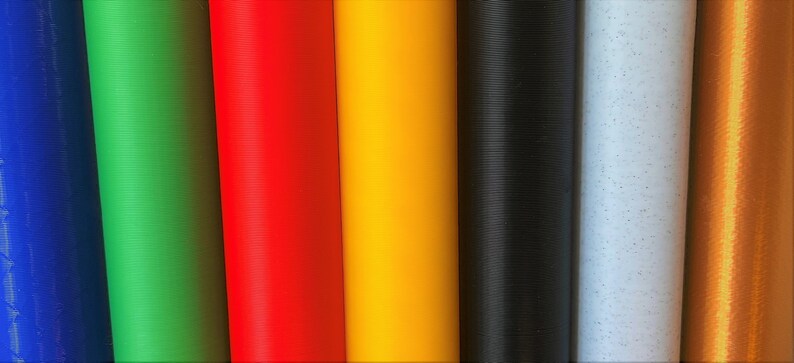 K&ouml;nnte beinhalten: Nahaufnahme von acht verschiedenen farbigen Filamentrollen f&uuml;r einen 3D-Drucker. Die Farben sind blau, gr&uuml;n, rot, orange, gelb, schwarz, wei&szlig; und kupfer.
