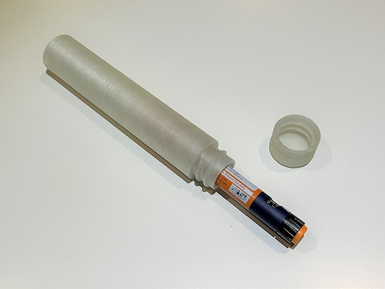K&ouml;nnte beinhalten: Ein wei&szlig;es Plastiketui mit Deckel, das f&uuml;r einen medizinischen Stift konzipiert ist. Der Stift ist teilweise in das Etui gesteckt, wobei die orange Spitze sichtbar ist.
