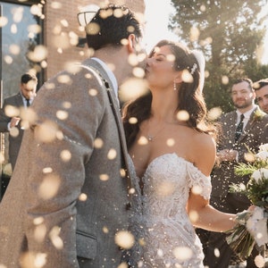 Peut inclure: Une mari&eacute;e et un mari&eacute; s'embrassent sous une pluie de confettis en quittant leur c&eacute;r&eacute;monie de mariage. La mari&eacute;e porte une robe blanche &agrave; &eacute;paules d&eacute;nud&eacute;es avec des d&eacute;tails en dentelle. Le mari&eacute; porte une veste de costume grise.