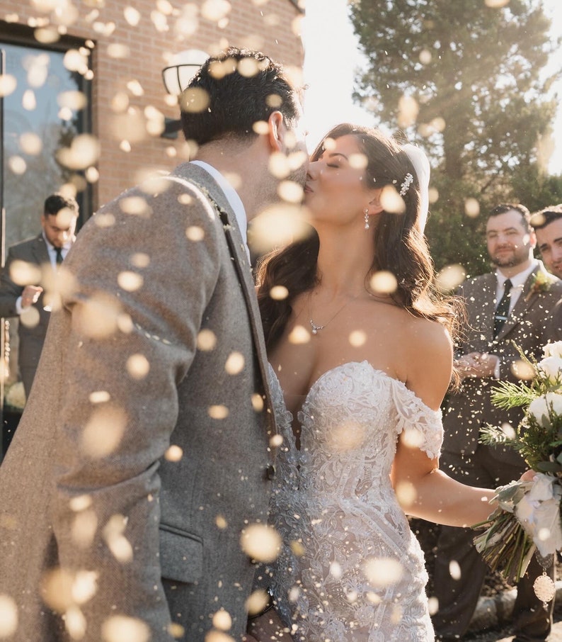 Peut inclure: Une mari&eacute;e et un mari&eacute; s'embrassent sous une pluie de confettis en quittant leur c&eacute;r&eacute;monie de mariage. La mari&eacute;e porte une robe blanche &agrave; &eacute;paules d&eacute;nud&eacute;es avec des d&eacute;tails en dentelle. Le mari&eacute; porte une veste de costume grise.